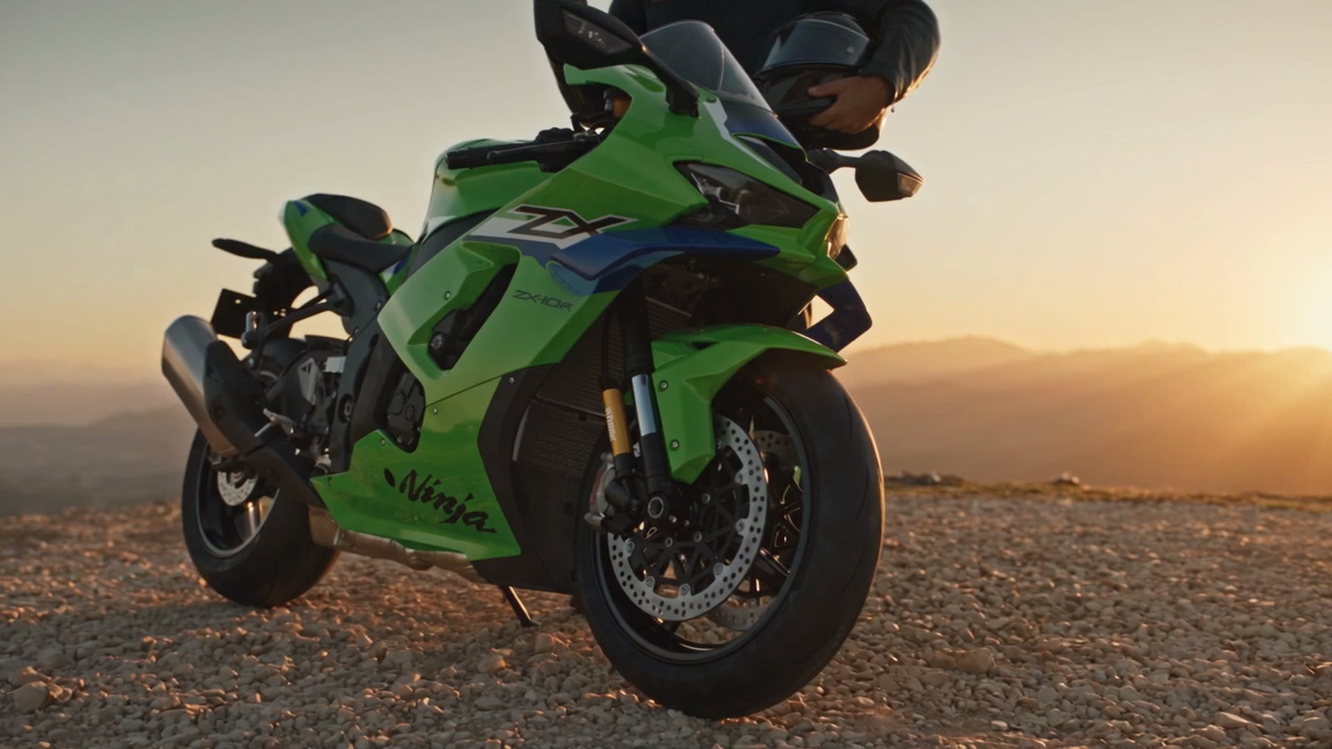 Una sportiva verde ferma su un piazzale di ghiaia al tramonto; dietro, in piedi, il motociclista guarda il telefono