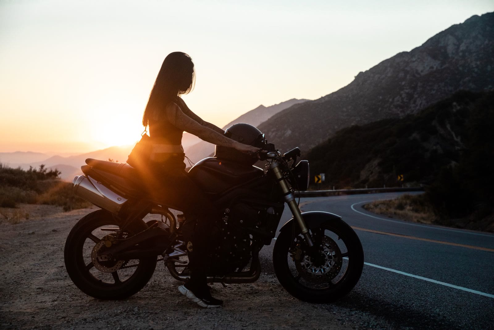 Una motociclista ferma in sella su una curva di montagna, controluce