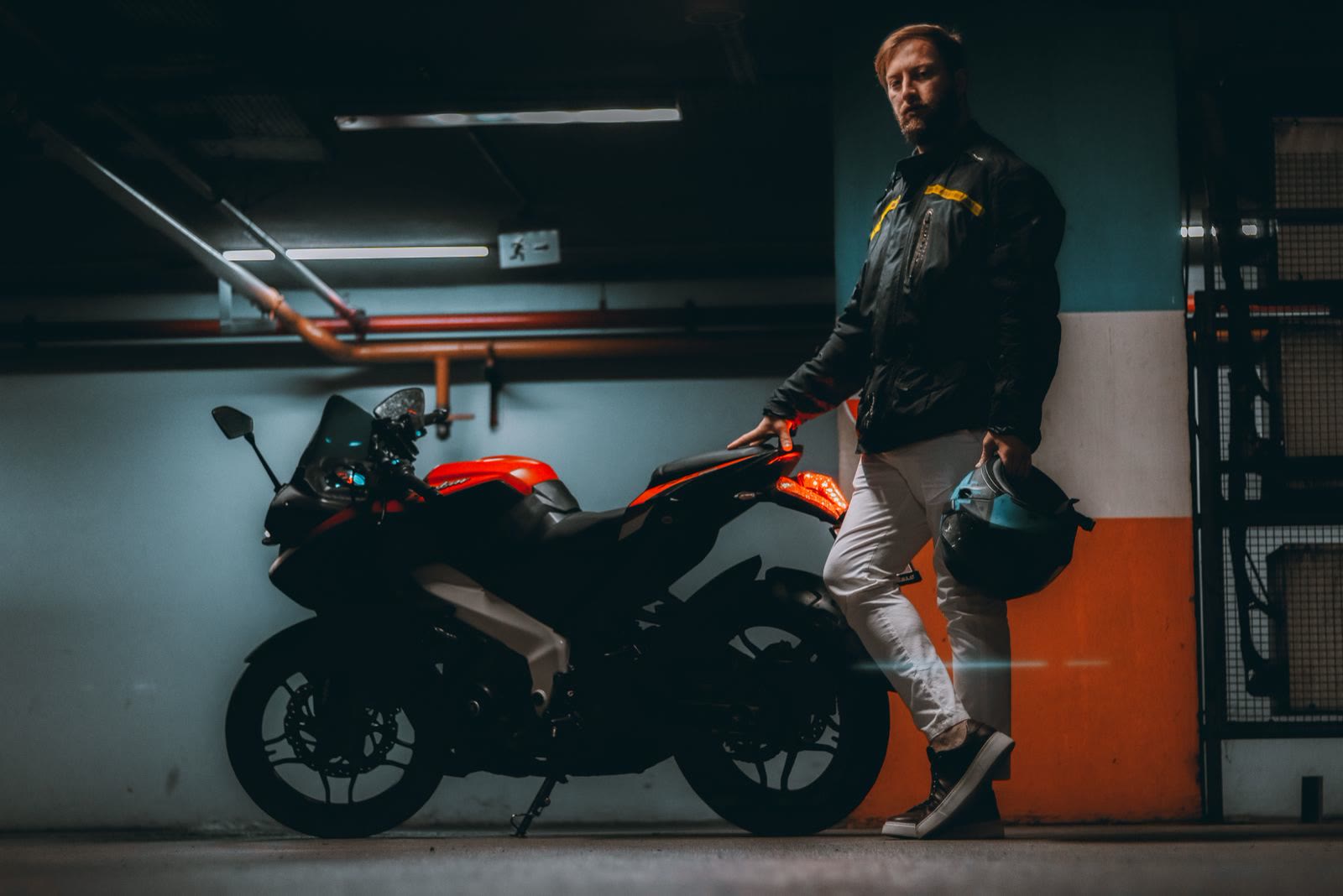 Un motociclista con il casco in mano accanto a una sportiva nera e arancio, in un garage