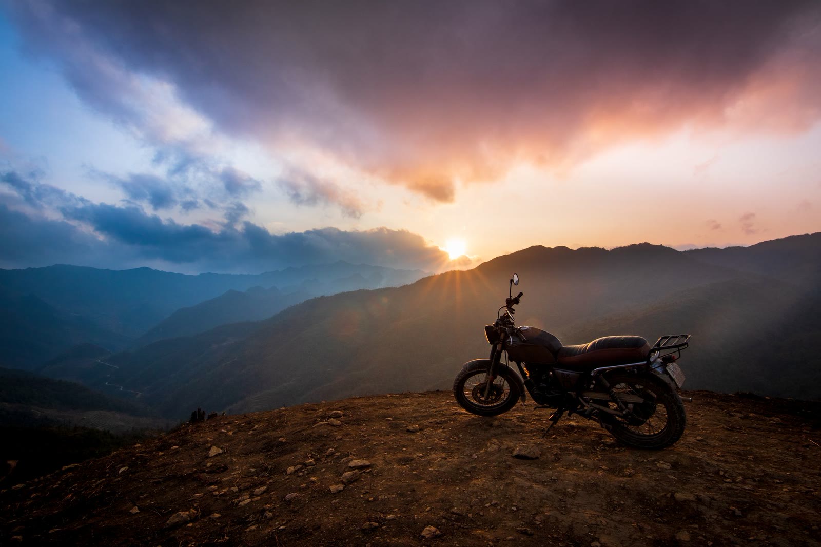 Una moto ferma su un crinale al tramonto, con le nuvole accese sopra una catena di montagne