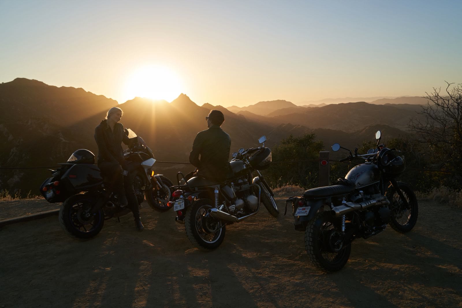 Due motociclisti e tre moto in controluce, fermi su un piazzale mentre il sole scende dietro le montagne
