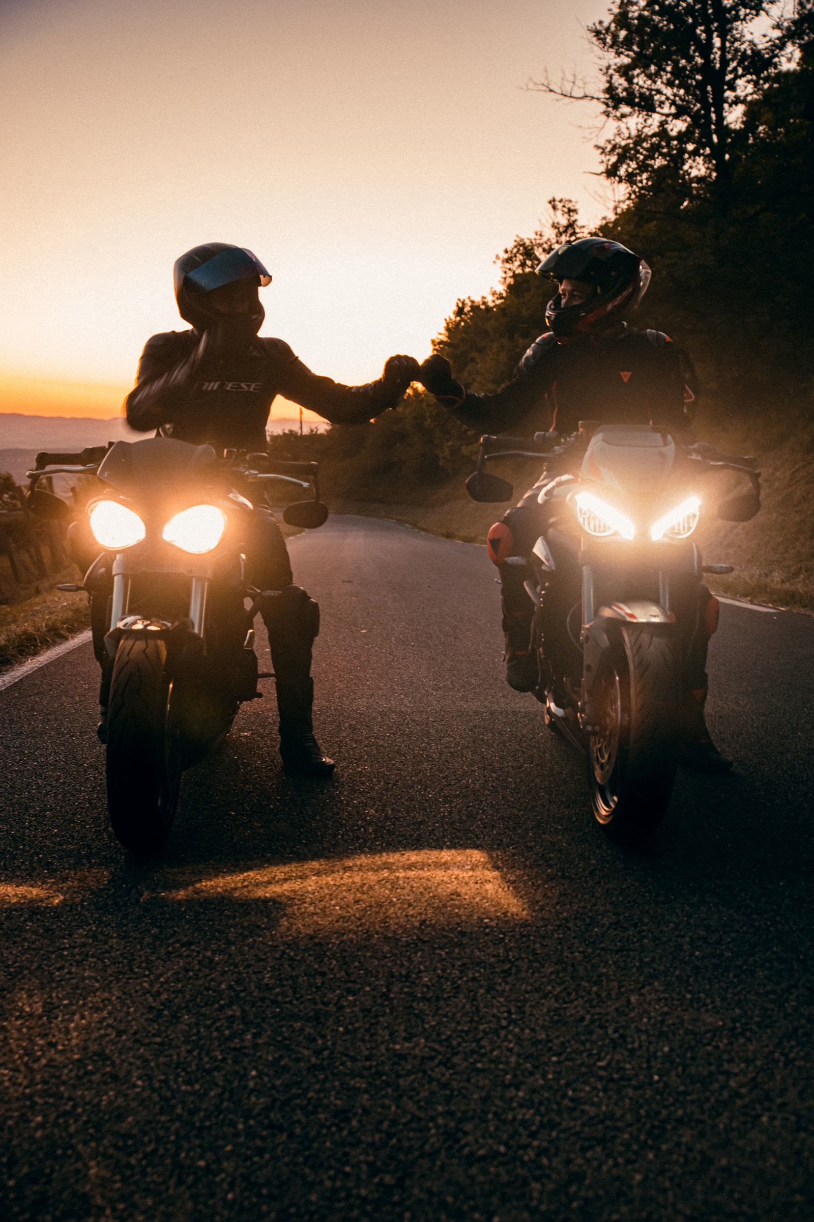 Due motociclisti affiancati si toccano il pugno al tramonto, con i fari accesi