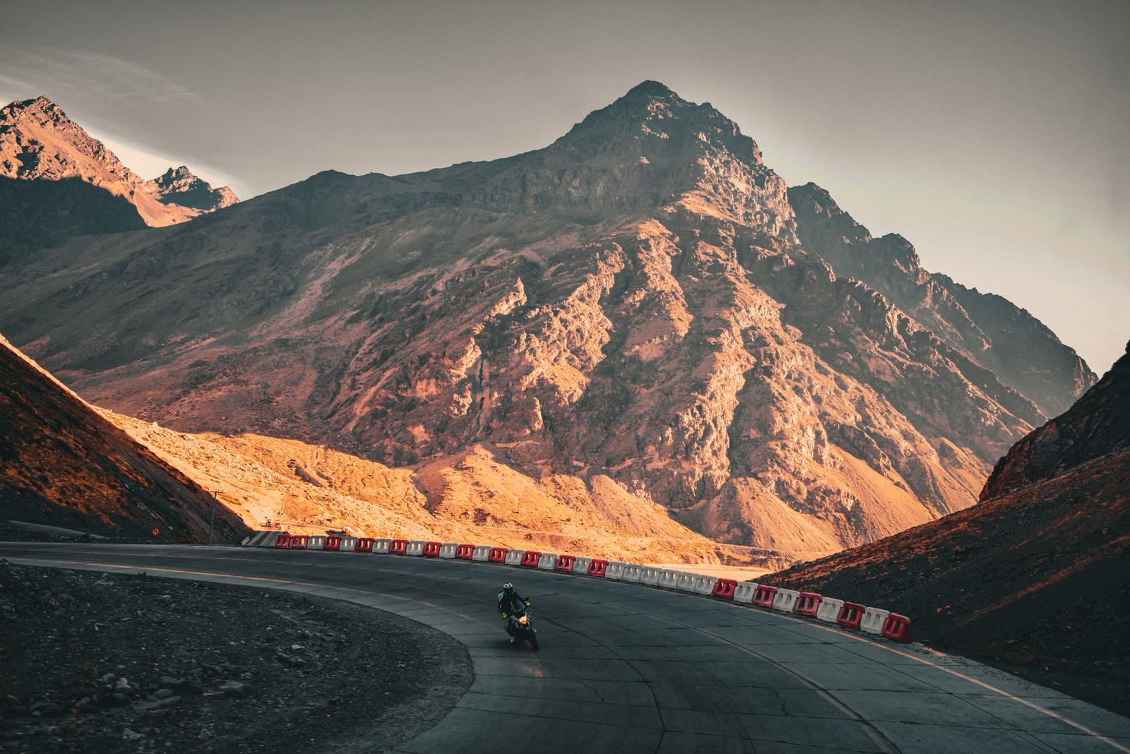 Una strada di montagna in curva con una moto sola, sotto una cima illuminata dal sole basso